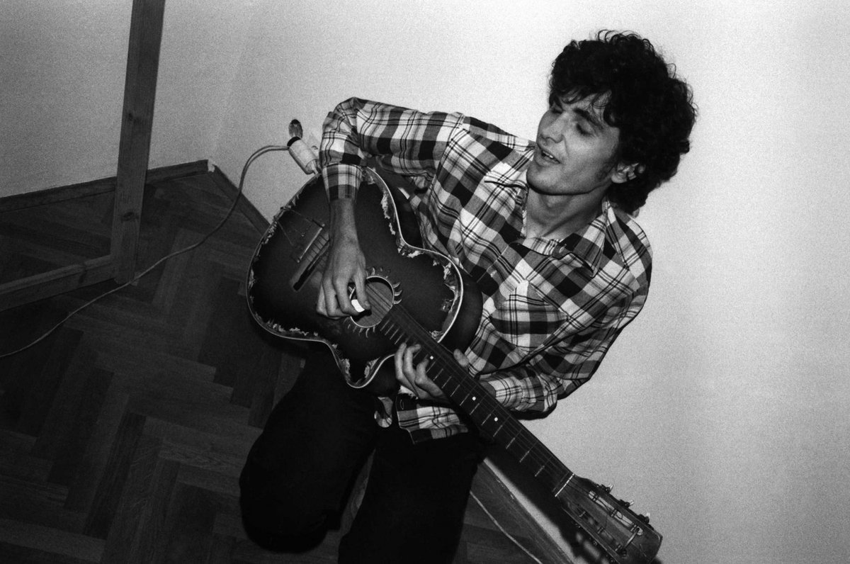 Young man in a plaid shirt playing a worn acoustic guitar indoors on a wooden floor.