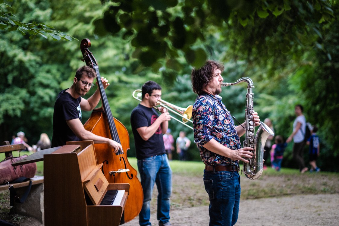 Outdoor park concert with a saxophonist, trombonist, and double bass player performing near a wooden piano while spectators watch.