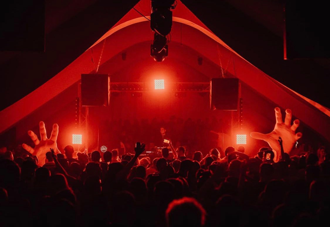 DJ on a red-lit stage with a dense crowd waving hands in the air at a nightclub or festival tent.