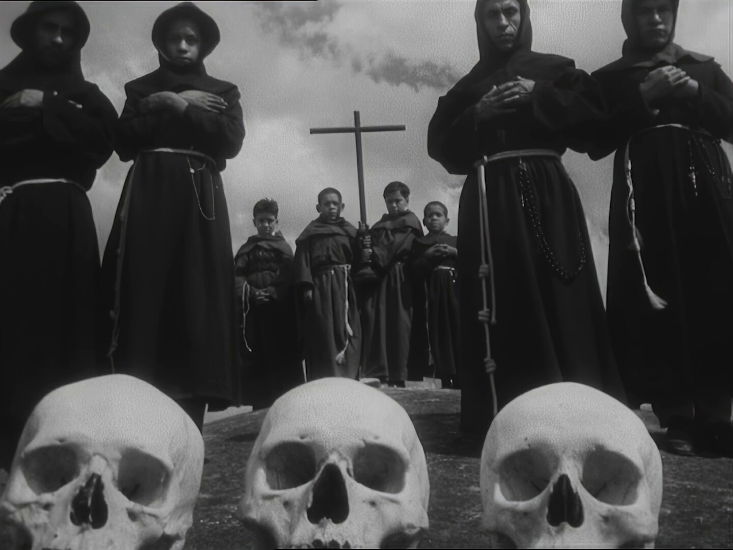 Hooded monks in dark robes stand with hands crossed around a central cross; foreground shows skulls, creating a grim, funeral scene.