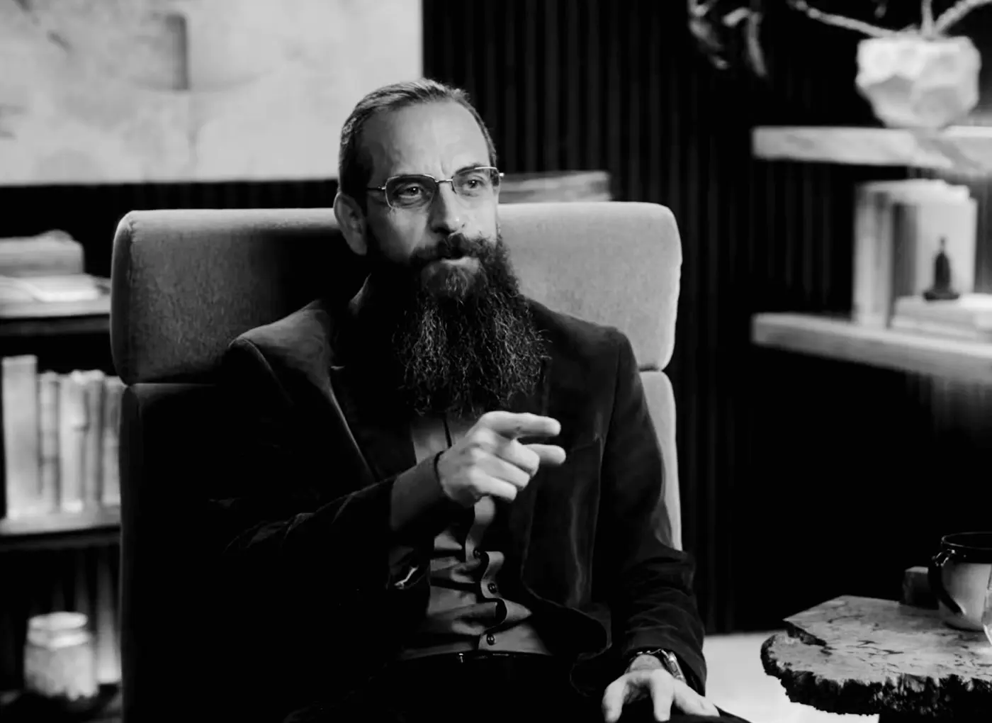 Man with glasses and a long beard sits in an armchair, gesturing with his hand as if speaking in a conversation.