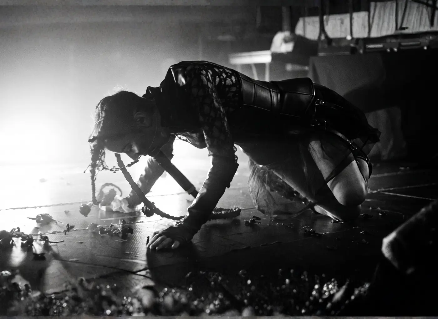 Crowded live stage with a performer on hands and knees, wearing a leather outfit, dramatic backlight highlighting their braided hair.