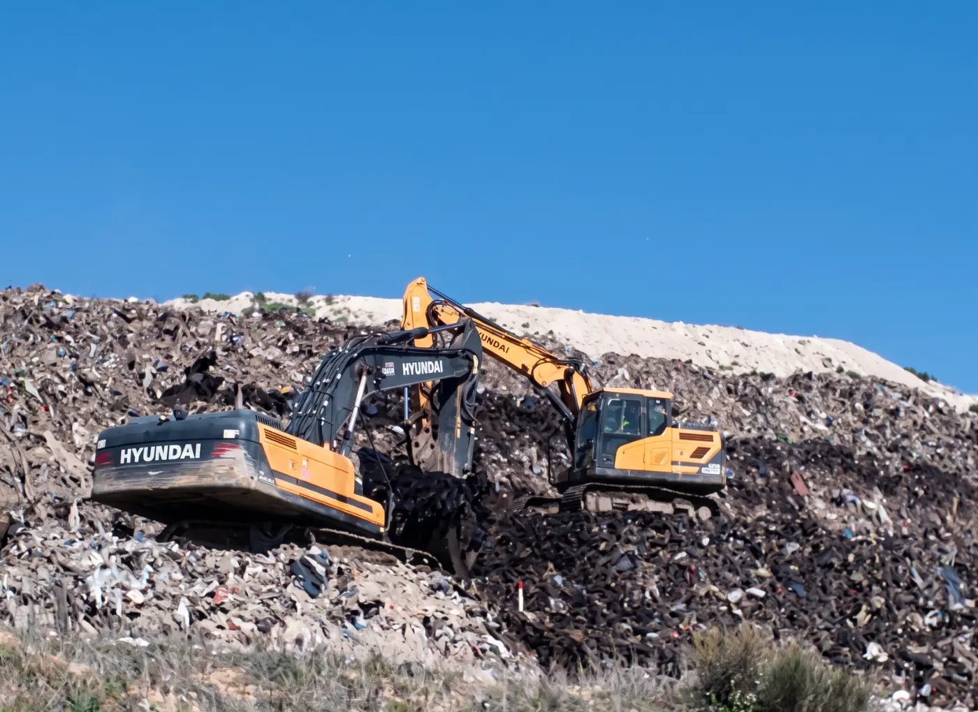 Two orange Hyundai excavators working on a landfill mound under a clear blue sky.