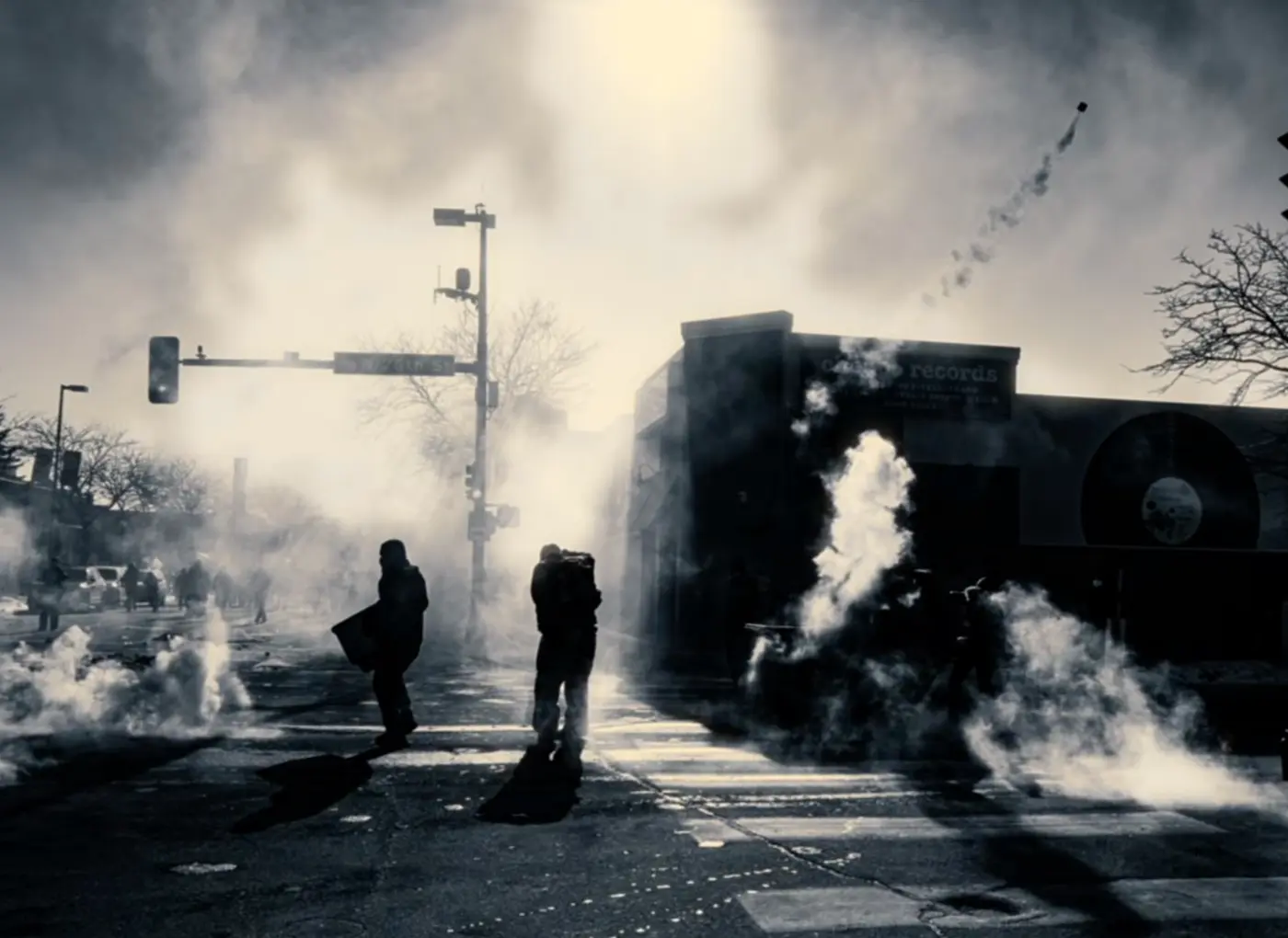 Protesters walk through a smoky urban street with crosswalk stripes and scattered debris, silhouettes visible through the haze.
