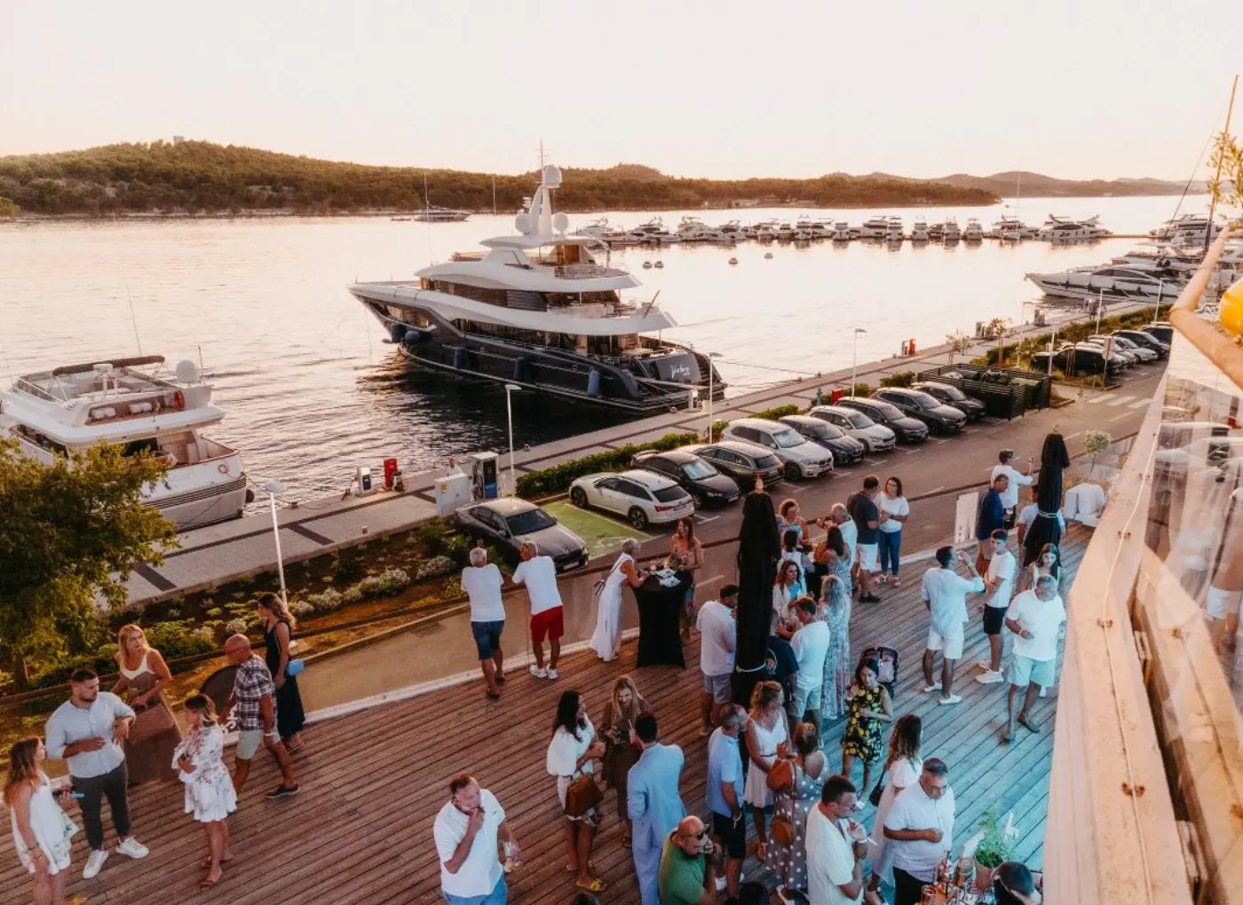 People mingling on a wooden marina deck at sunset, with luxury yachts anchored in calm water behind them.