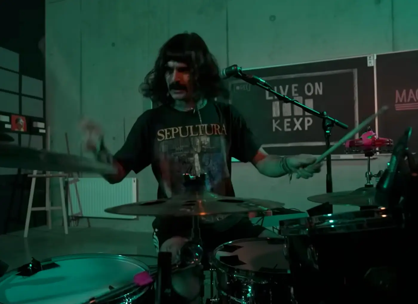 Drummer with long hair wearing a Sepultura shirt plays a drum kit in a studio under green lighting, with a 'Live on KEXP' sign in the background.