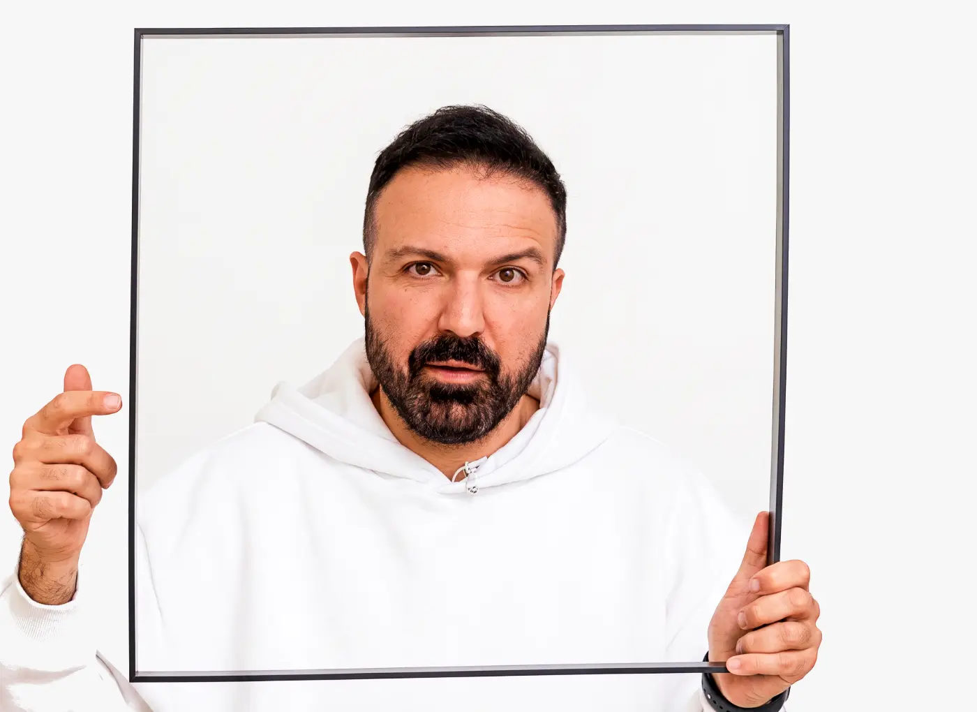 Portrait of a bearded man in a white hoodie holding a black square frame around his upper body against a white background, facing the camera.