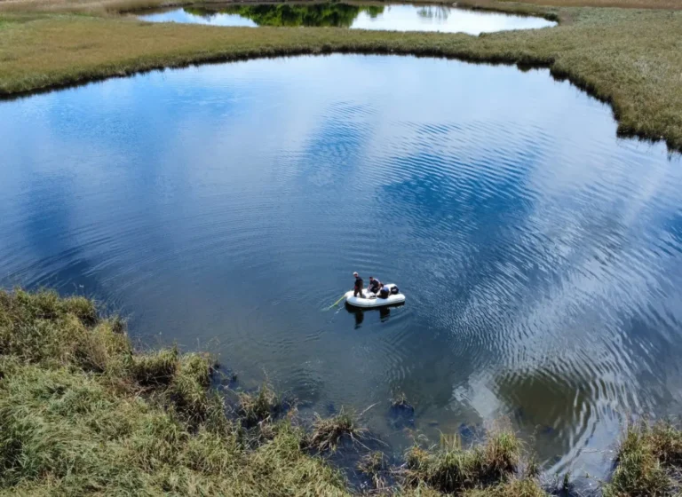 A small inflatable raft with four people paddling on a calm, circular blue pond surrounded by grassy banks.