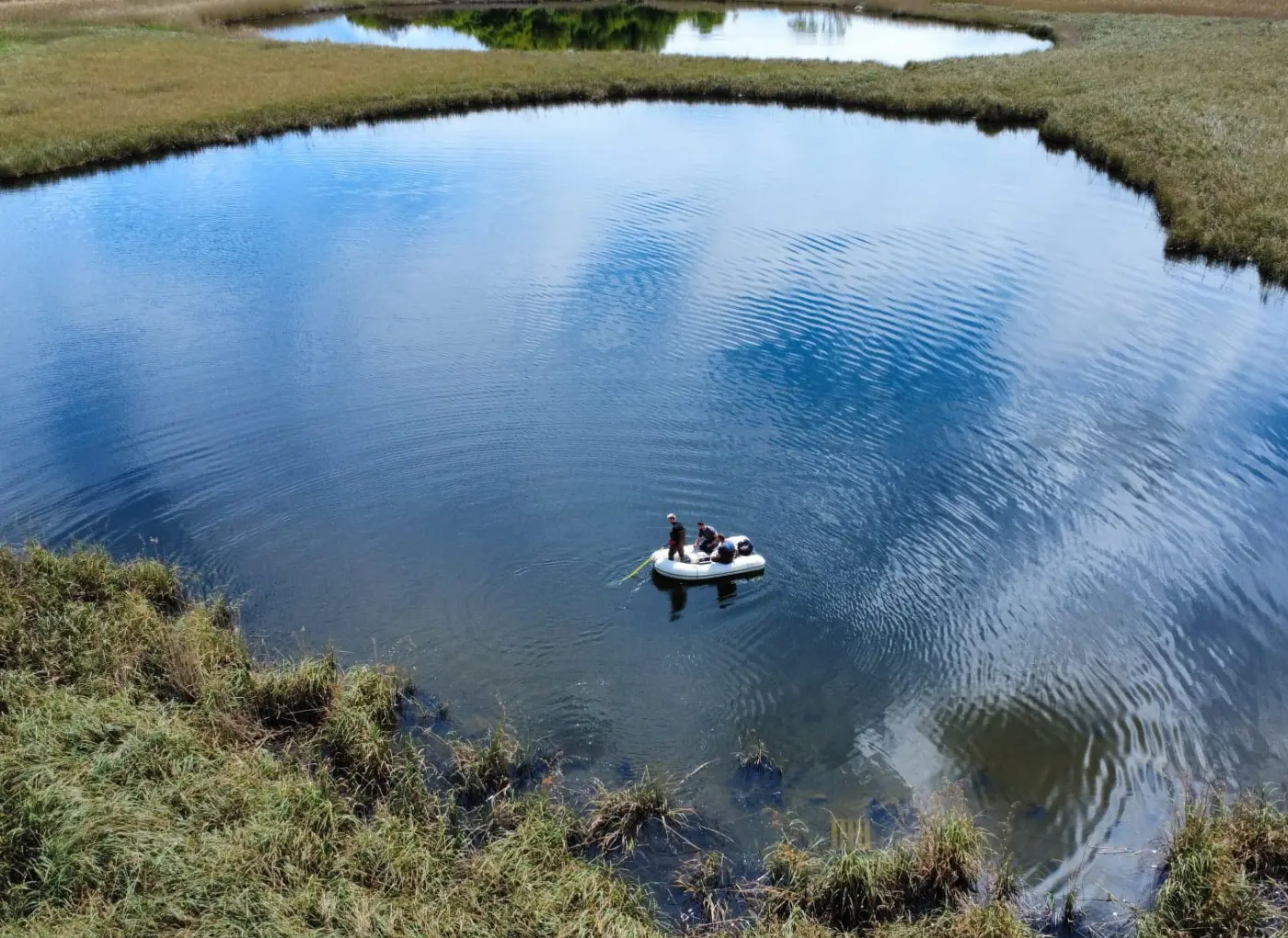 A small inflatable raft with four people paddling on a calm, circular blue pond surrounded by grassy banks.