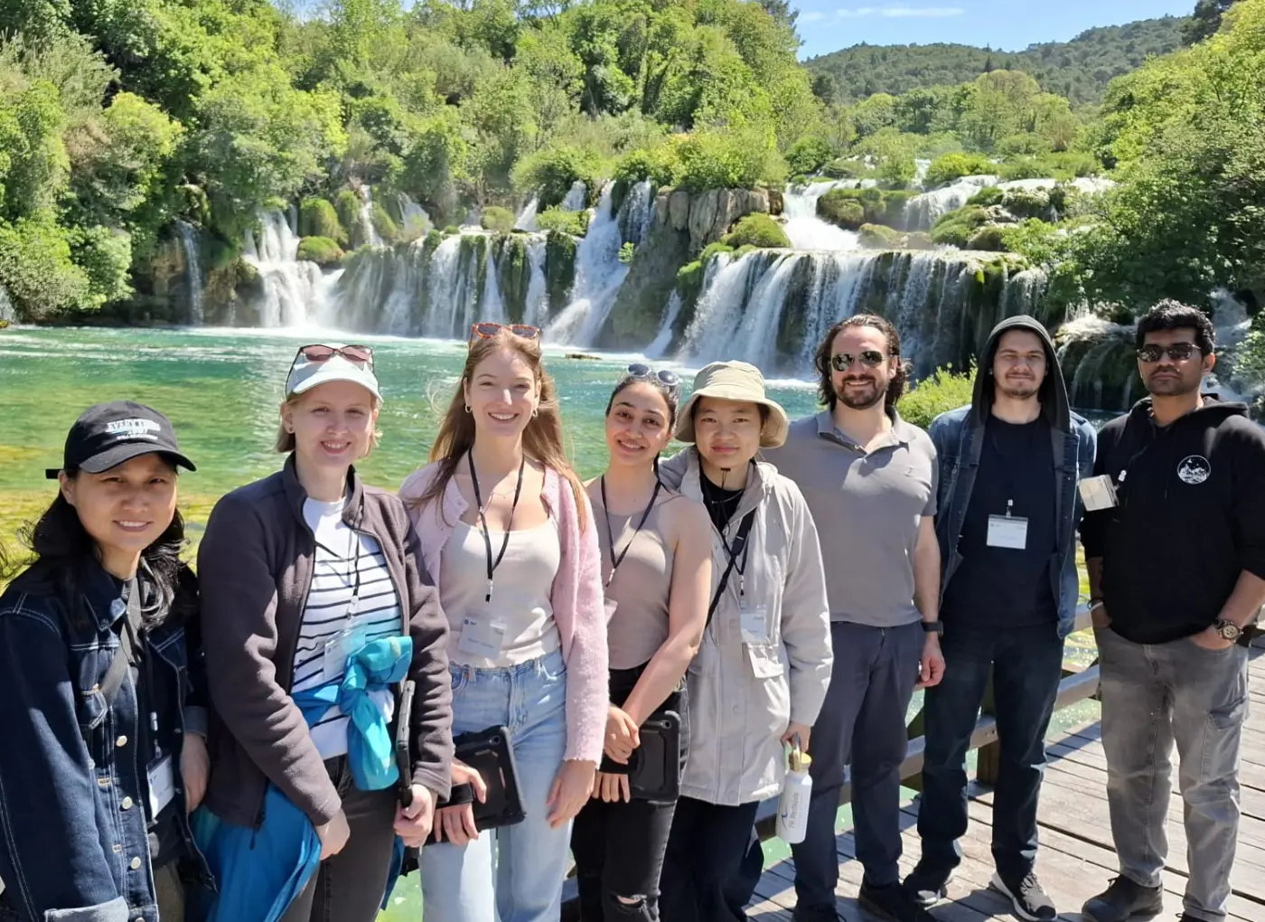 Nine people smiling on a wooden boardwalk with a multi-tier waterfall and green forest behind them, sunny day by the waterway.
