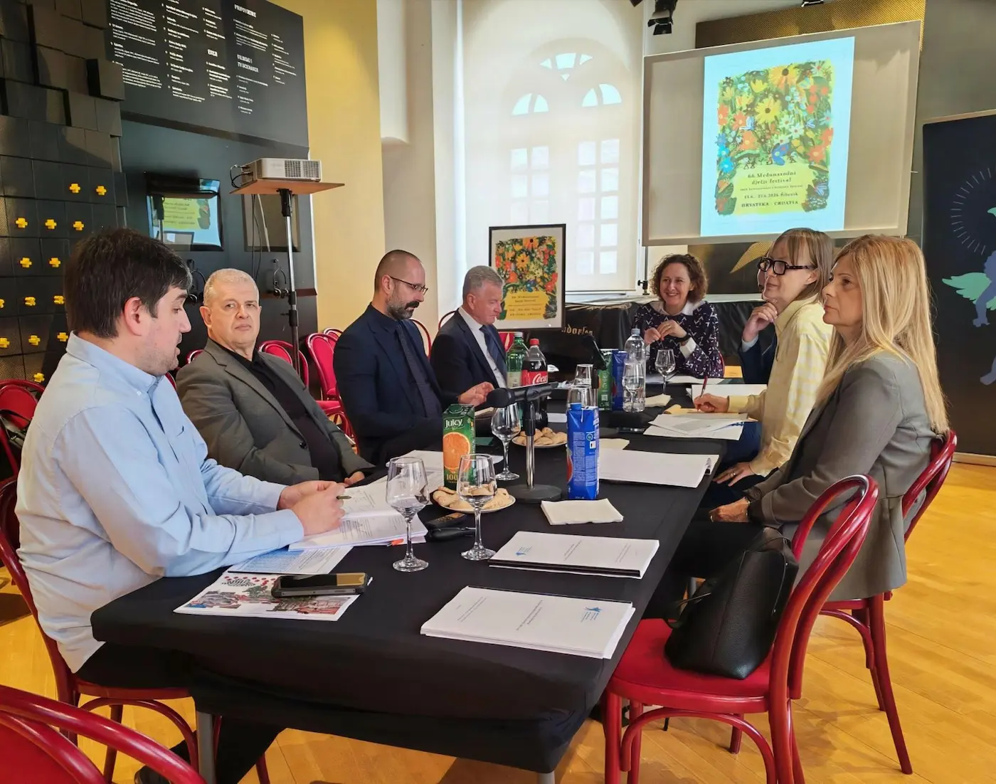 Panel of six people seated at a long table with papers, drinks, and microphones during a meeting in a bright room with a projected poster on the screen.
