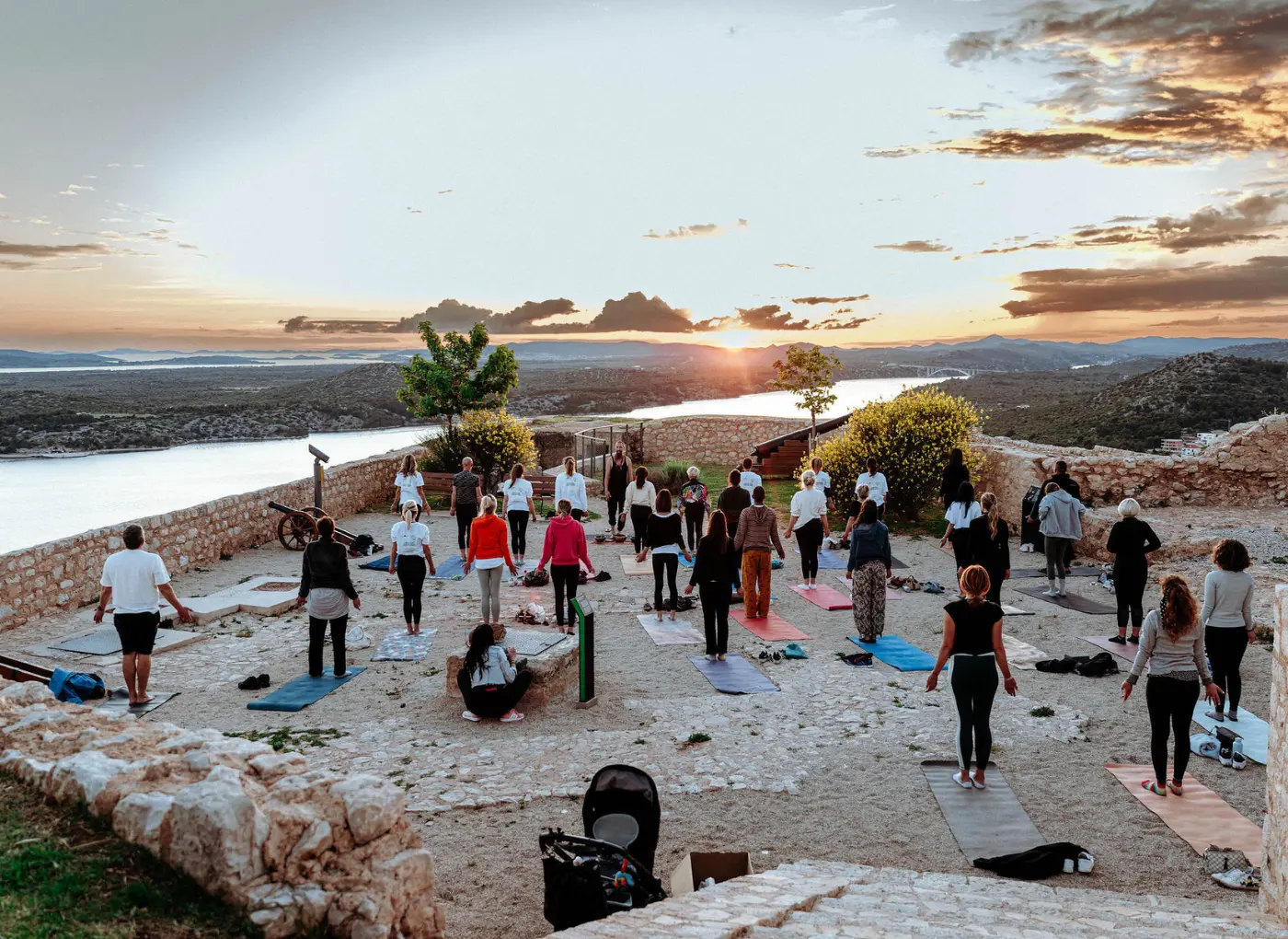Group of people practicing outdoor yoga on mats at sunset overlooking a river valley.