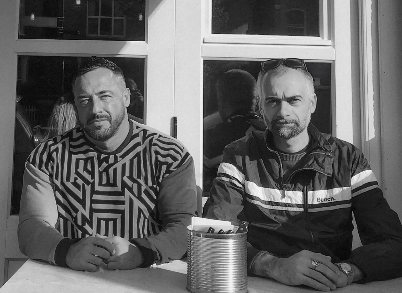 Two men sit side by side at a table outside a building, facing the camera; left man wears a geometric patterned sweatshirt, right man wears a striped jacket with sunglasses on his head on top of his head.