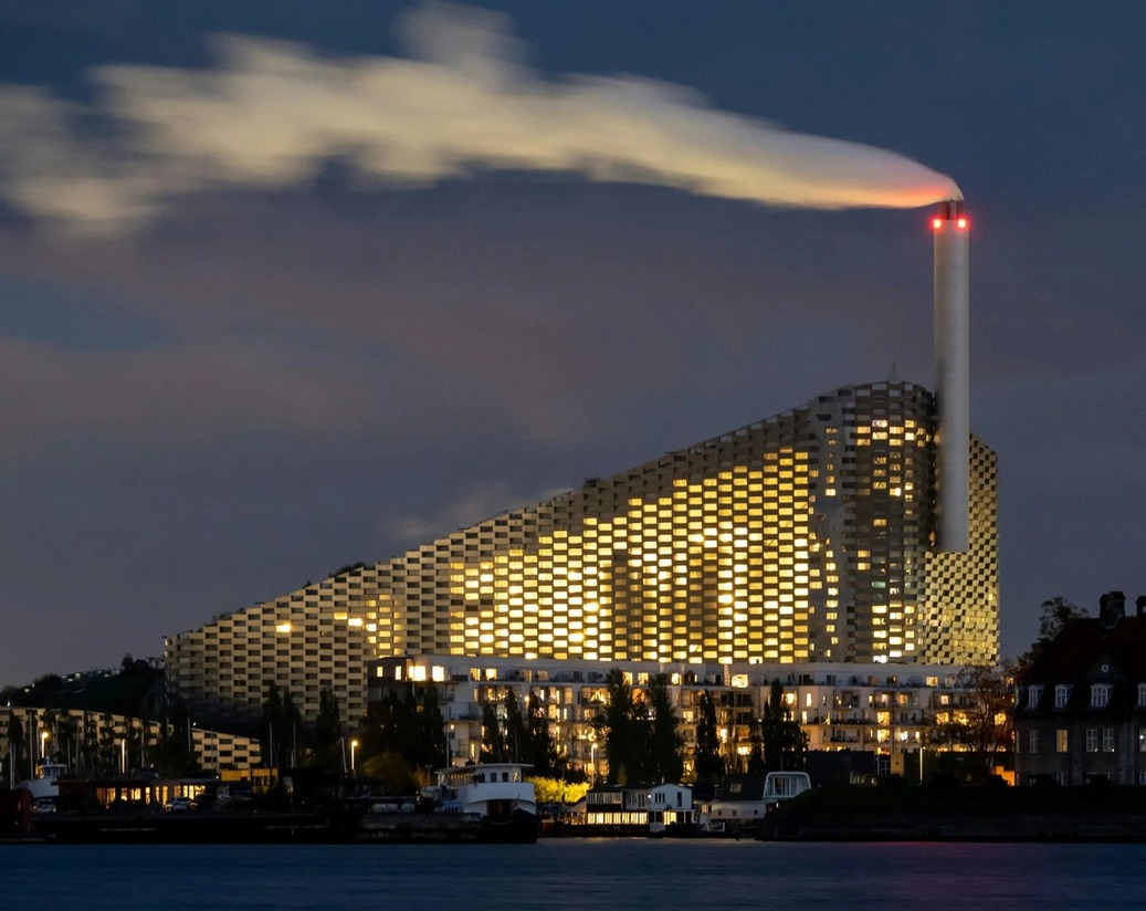 Nighttime view of a futuristic building with a slanted, grid-like facade lit from within and a tall smokestack emitting smoke.