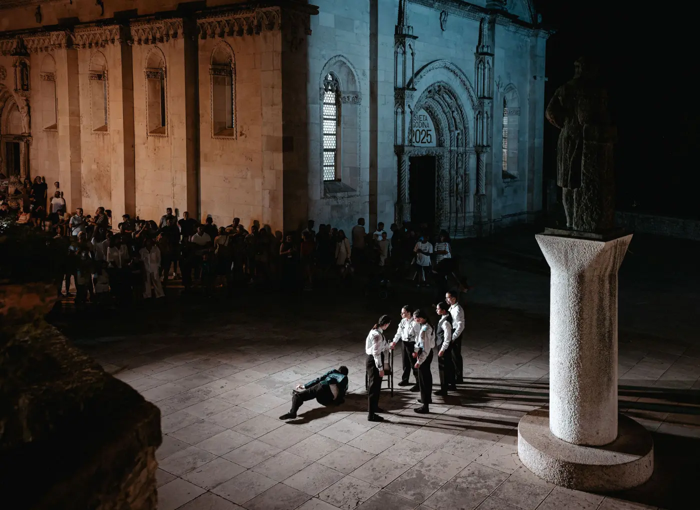 Night outdoor performance outside a historic stone church; six performers in white shirts and black pants stand around a person on the ground while a crowd watches.