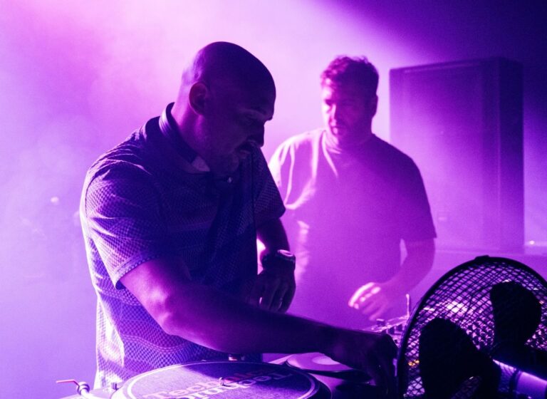 DJ wearing headphones scratching vinyl on a turntable while another DJ watches in a purple-lit club over smoky air