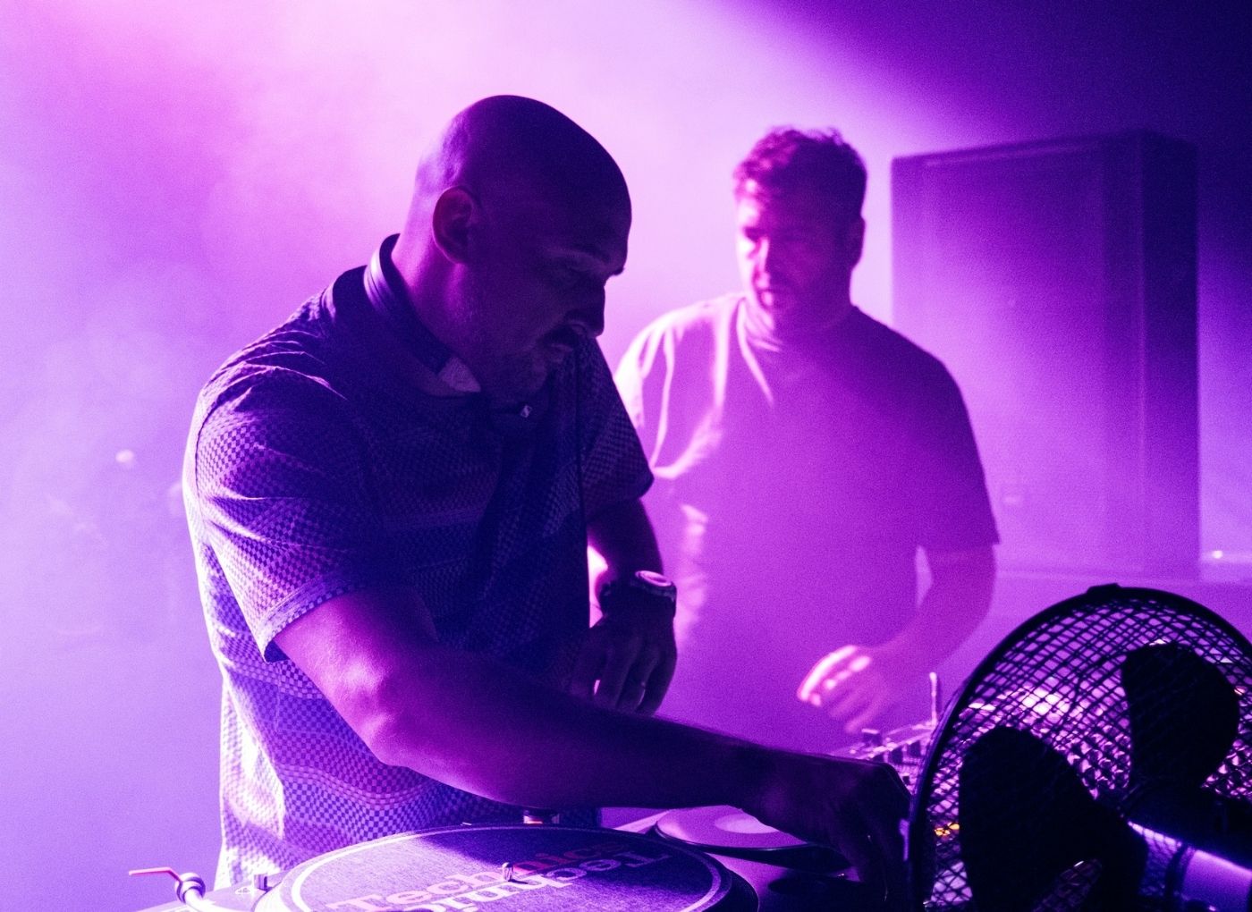 DJ wearing headphones scratching vinyl on a turntable while another DJ watches in a purple-lit club over smoky air
