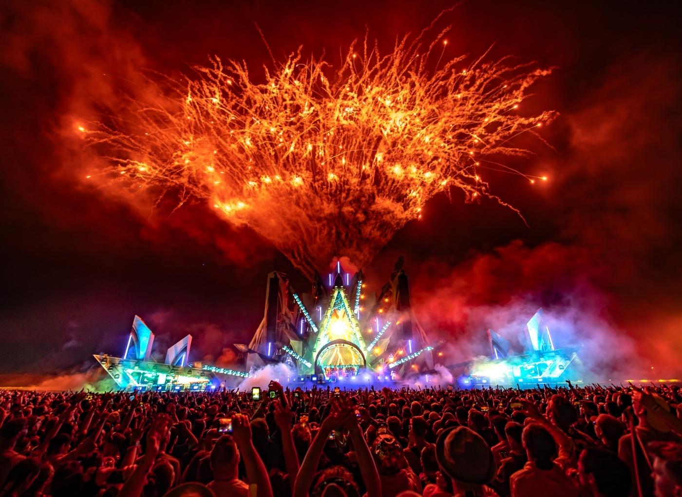 Thrilling outdoor concert with a neon-lit stage and a massive cheering crowd as fireworks explode overhead at night.