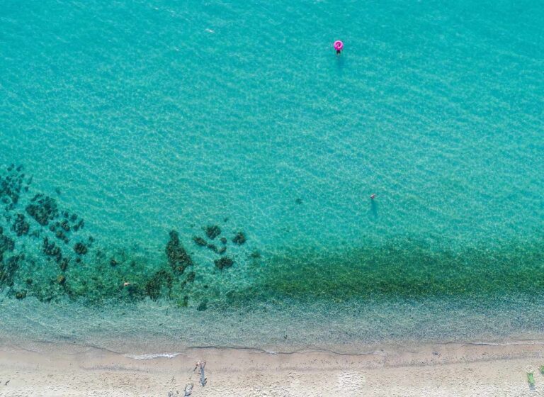 Aerial view of clear turquoise sea along a sandy beach with two swimmers and a pink inflatable ring near the surface.