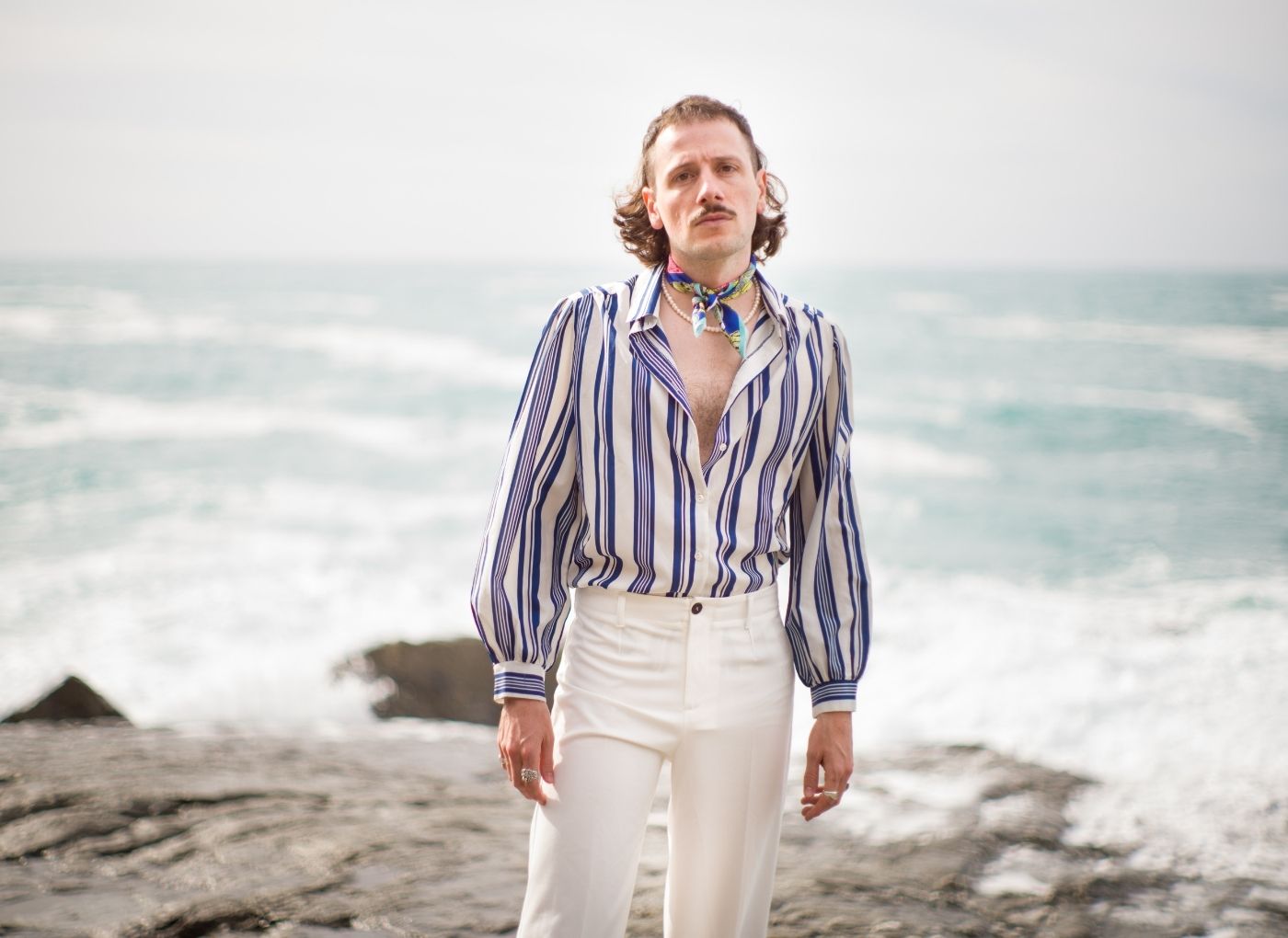 Man standing on a rocky beach by the sea, wearing a blue-and-white striped shirt, white pants, and a colorful scarf at the neck.