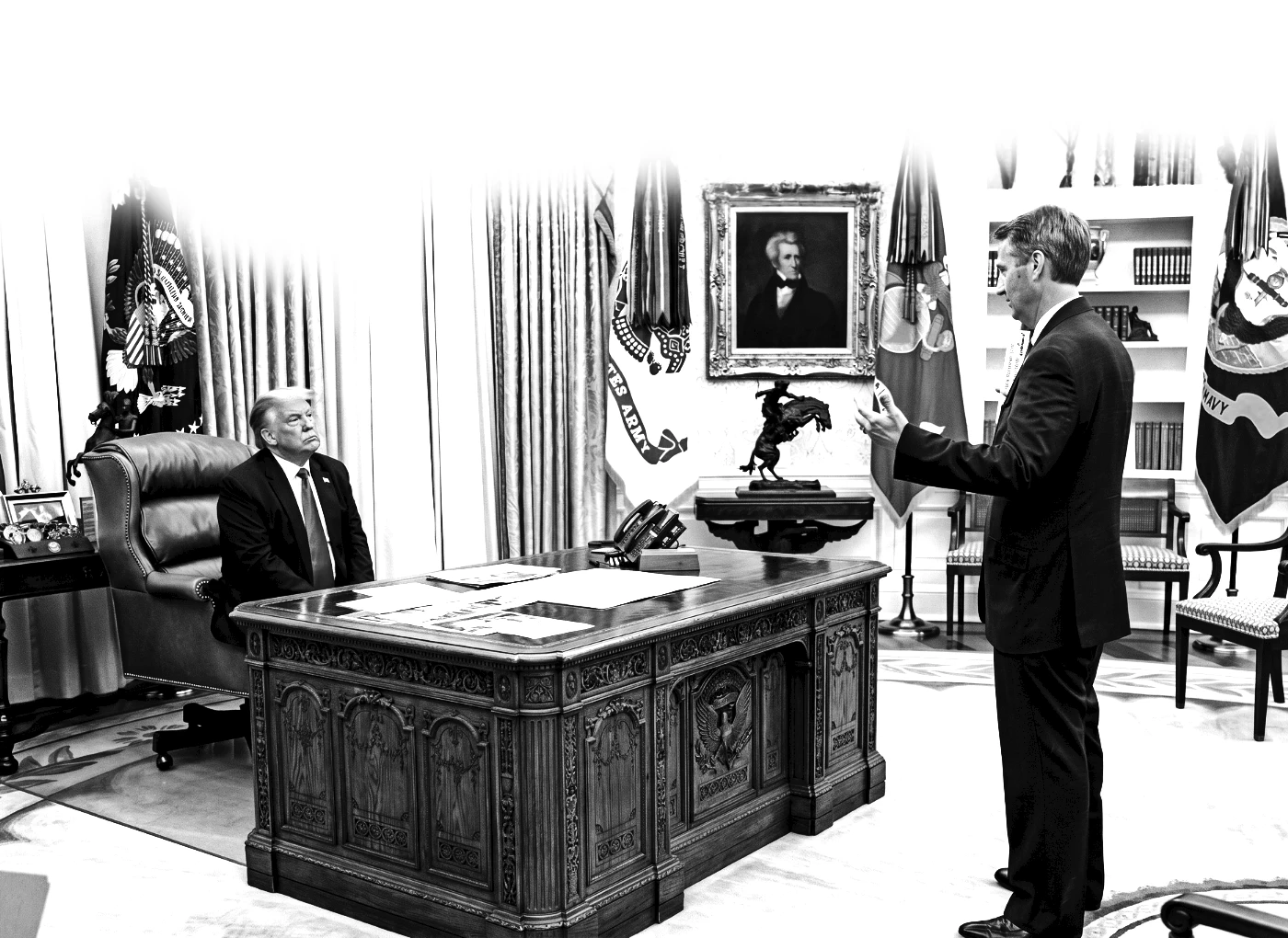 Two men in suits in a formal government office: one seated behind an ornate carved desk, the other standing and speaking to him, with flags and a portrait in the background.