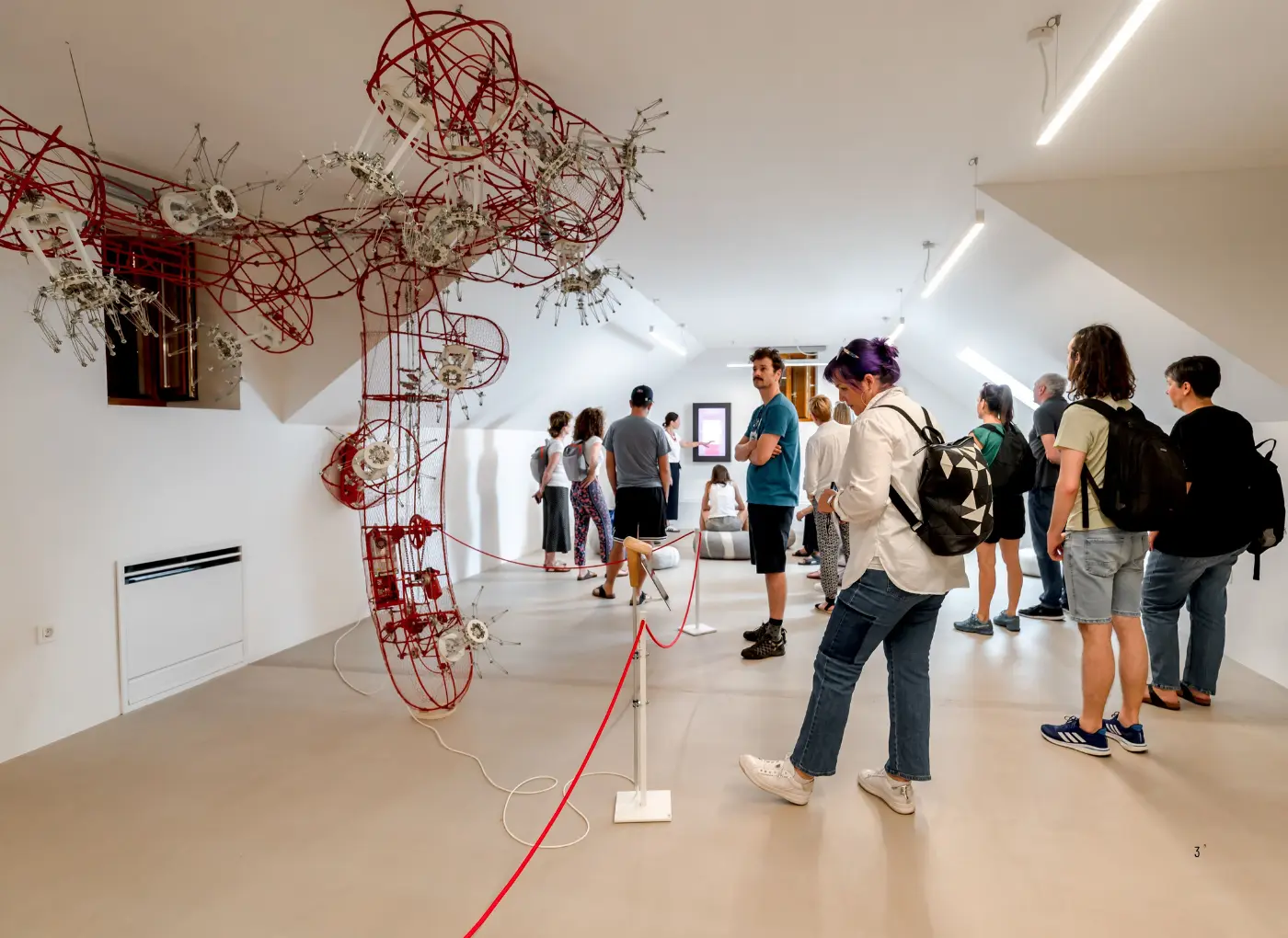 Gallery visitors observe a large red wire sculpture with gears and metal components suspended from the ceiling in a bright white room.