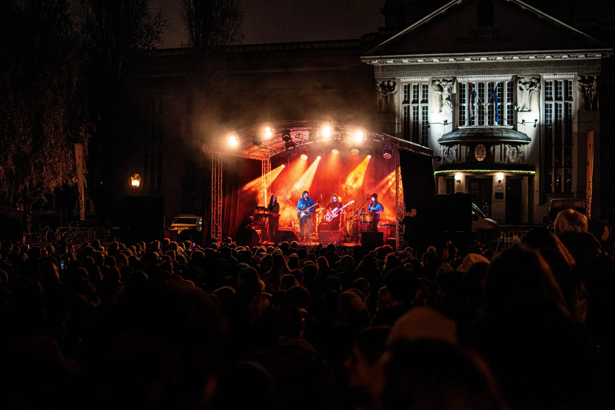 Outdoor nighttime concert with a band performing on a brightly lit stage in front of a historic building, crowd silhouettes visible in the foreground.