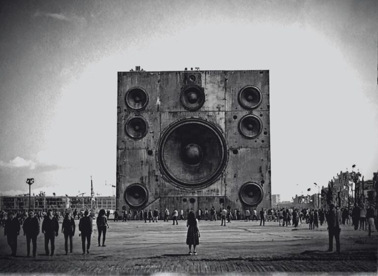 A colossal speaker sculpture with a central large woofer and multiple surrounding drivers in a wide plaza filled with people.