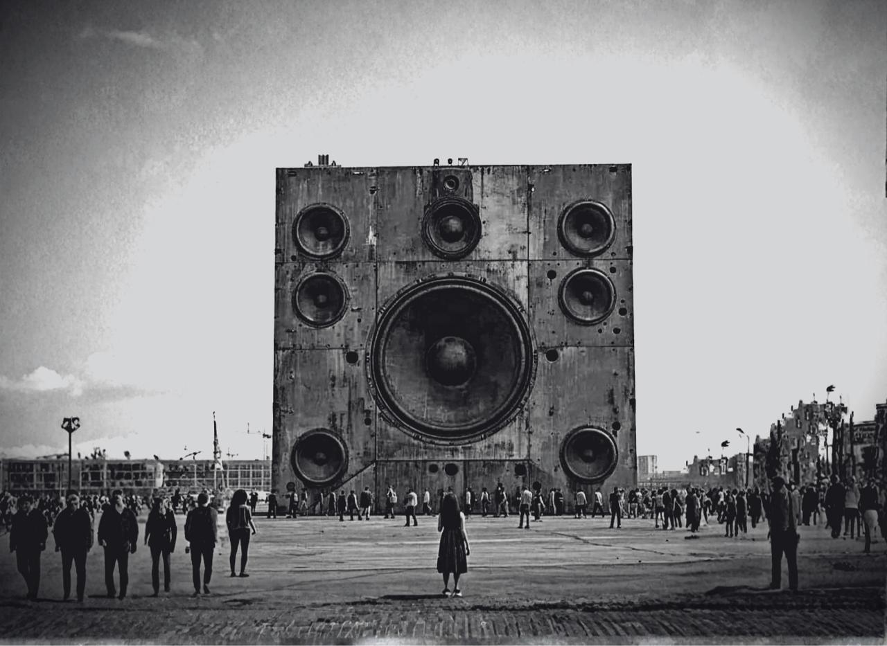 A colossal speaker sculpture with a central large woofer and multiple surrounding drivers in a wide plaza filled with people.