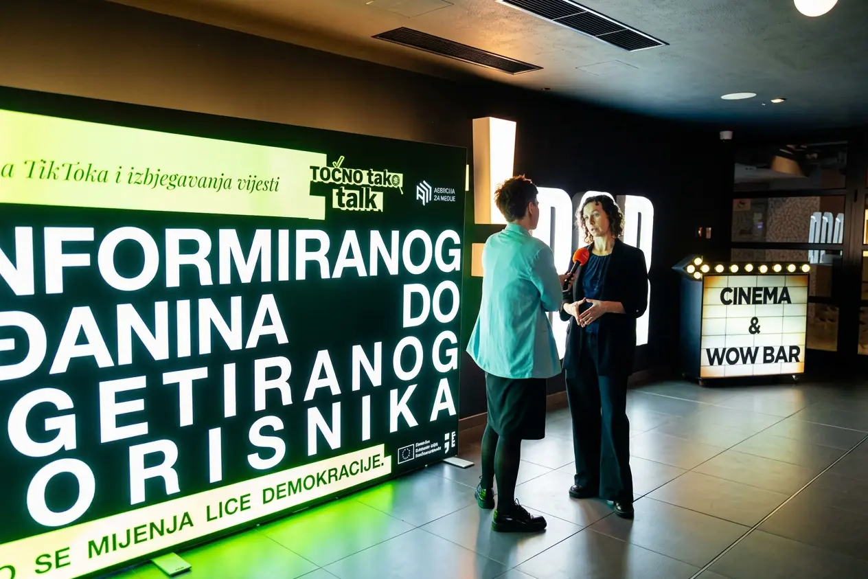 Two people converse in a cinema lobby, one holding an orange microphone, beside a bright green information board and a 'CINEMA & WOW BAR' sign.
