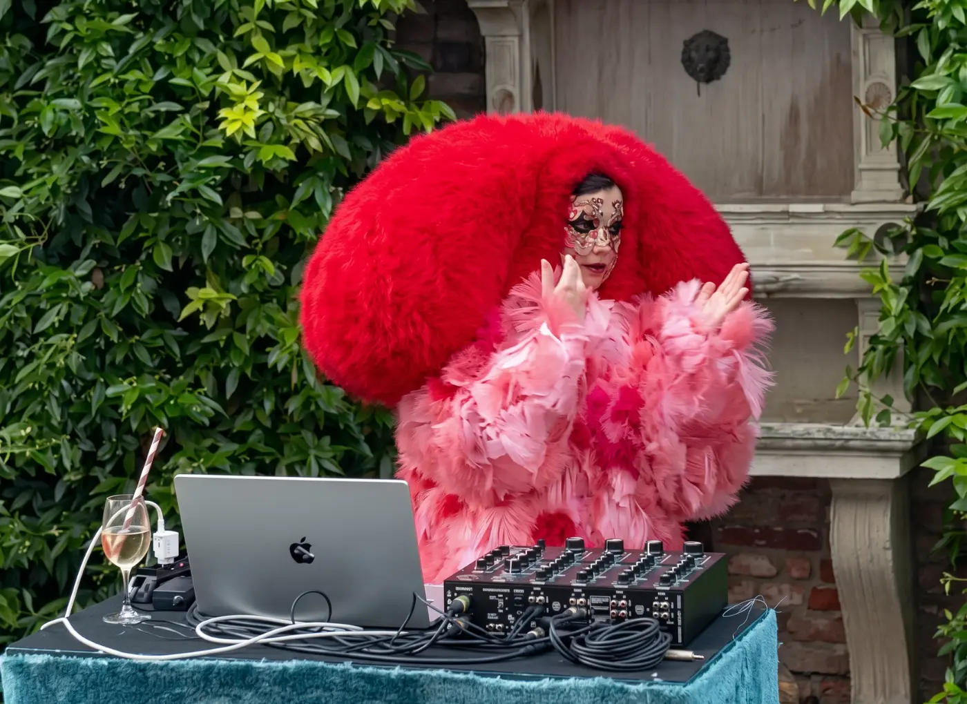 Person wearing a red fluffy headpiece and pink feathered coat performing as a DJ with laptop and mixer outdoors.