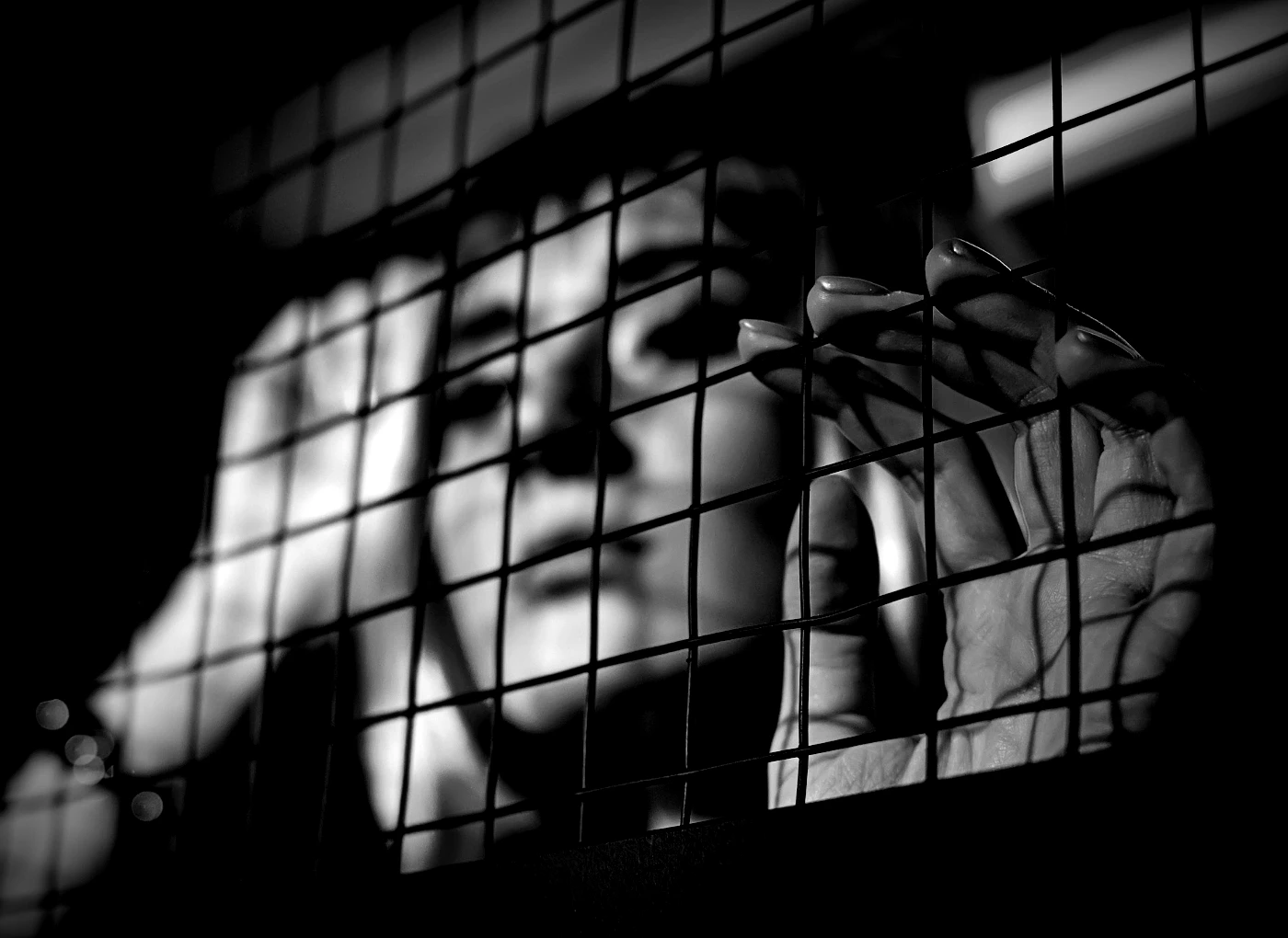 A hand grips a metal wire fence in black and white, with a blurred face visible behind the bars.