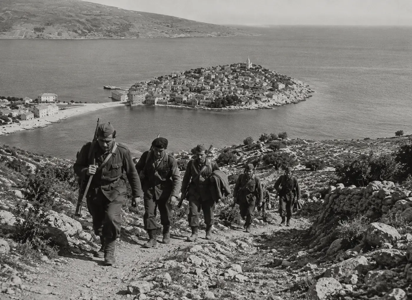 Soldiers in WWII uniforms march uphill along a rocky path toward a coastal village on a small island, with rifles slung and a calm sea in the background.