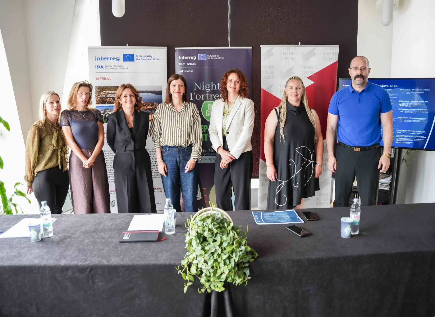 Seven professionals pose for a group photo behind a long black table with water bottles, banners, and a display screen in a bright conference room.