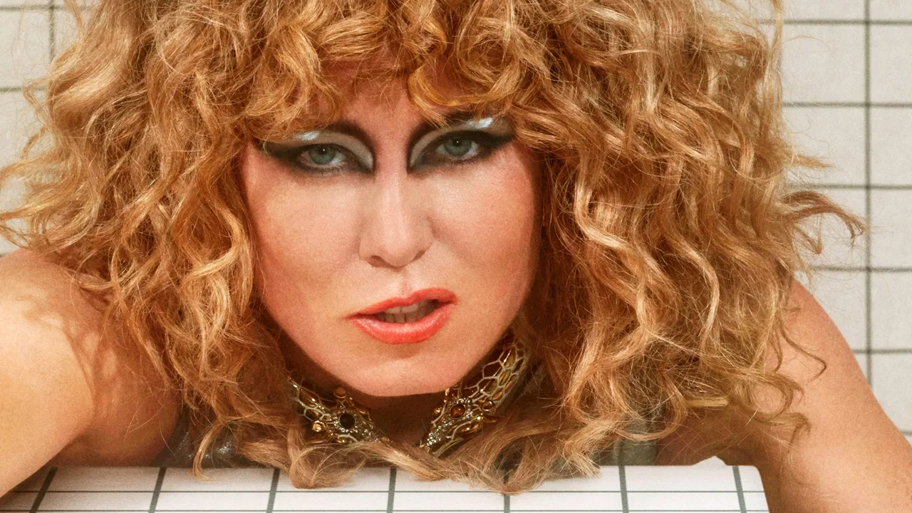 Close-up portrait of a woman with voluminous curly red hair, bold eye makeup, and coral lipstick, wearing a jeweled necklace against a tiled background.