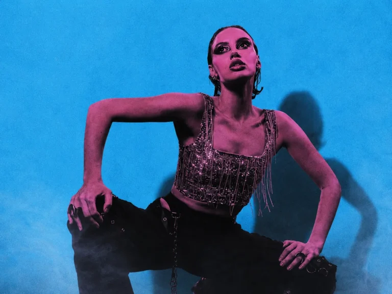 Model in a fringed sequined top crouches against a bright blue backdrop, pink lighting for a bold fashion shot.