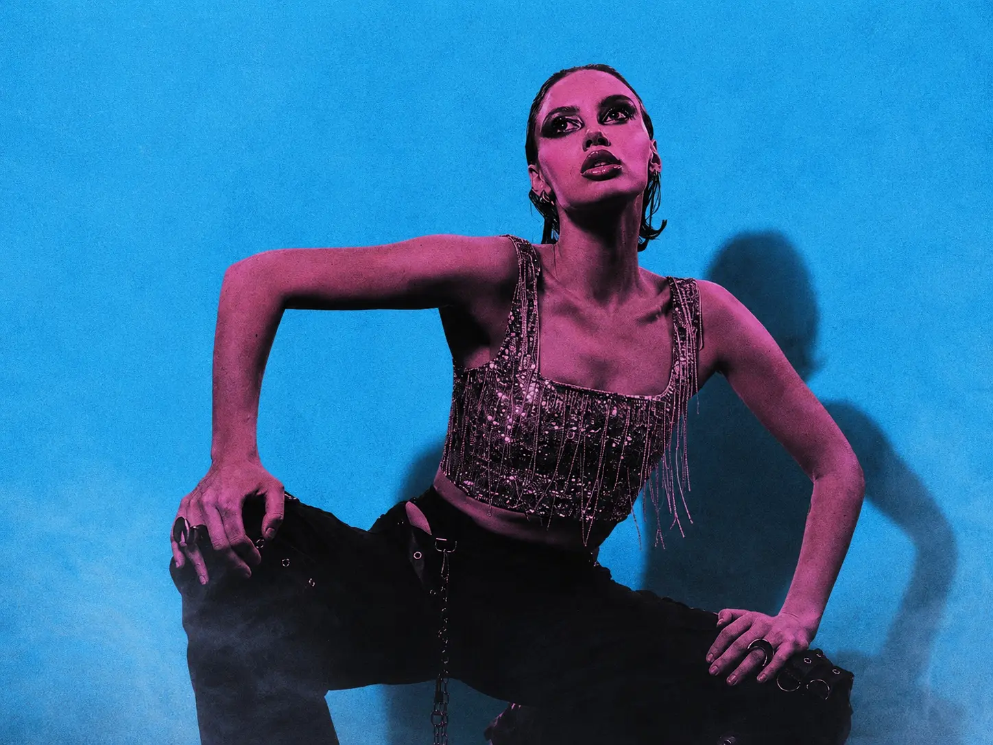 Model in a fringed sequined top crouches against a bright blue backdrop, pink lighting for a bold fashion shot.