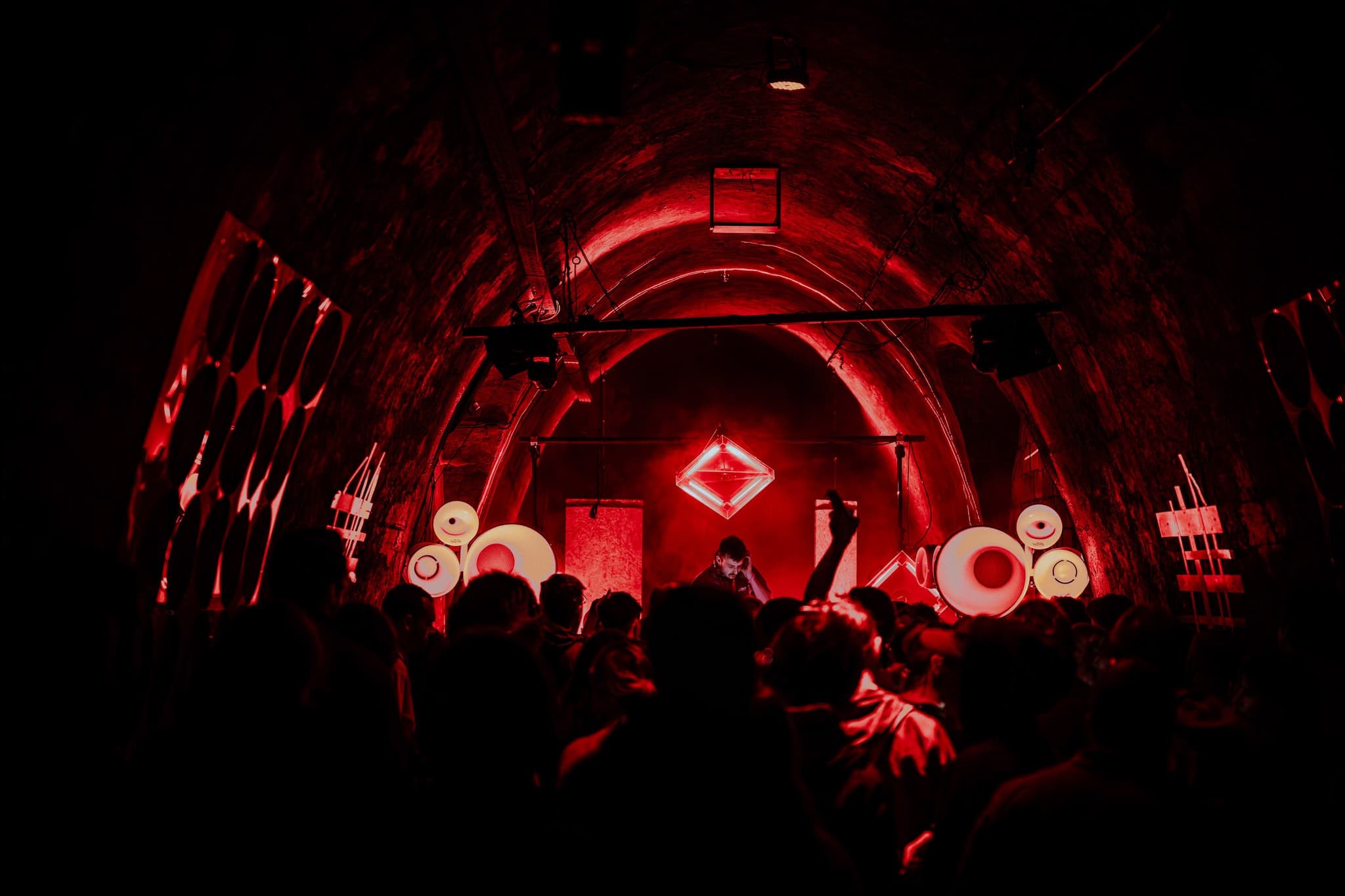 Dimly lit underground club with red lighting, curved brick arches, and a crowd dancing near neon geometric light fixtures.