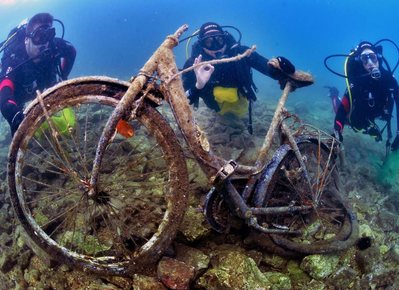 Three scuba divers hover over a rusted, sunken bicycle on a rocky seabed under clear blue water.
