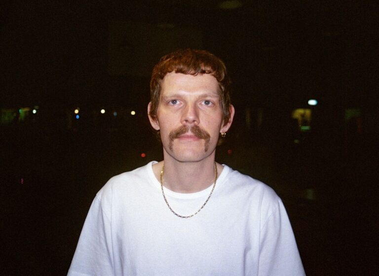 Portrait of a man with red hair and a mustache in a white T‑shirt and chain, standing at night with distant lights in the background