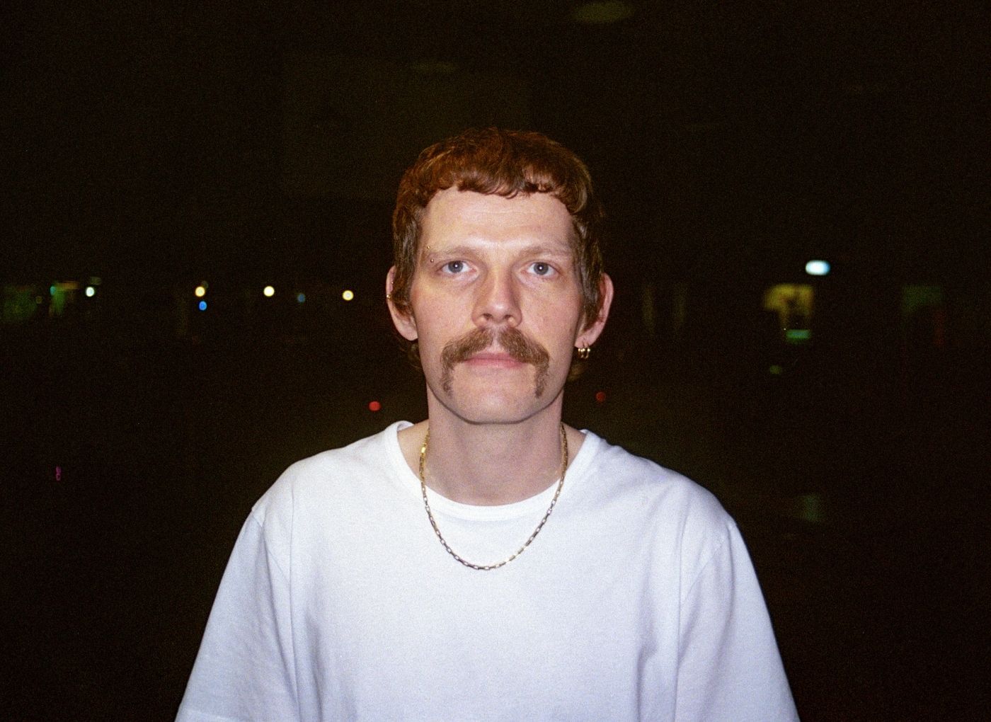Portrait of a man with red hair and a mustache in a white T‑shirt and chain, standing at night with distant lights in the background