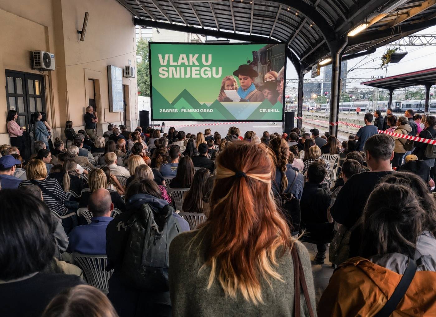 Large audience seated under a covered platform watching a film on a big screen with a green banner reading 'VLAK U SNIJEGU'.