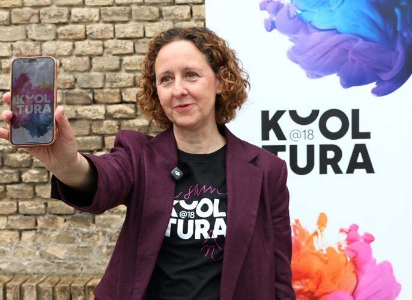 Woman in a burgundy blazer takes a selfie with her smartphone against a brick wall and a colorful event banner.