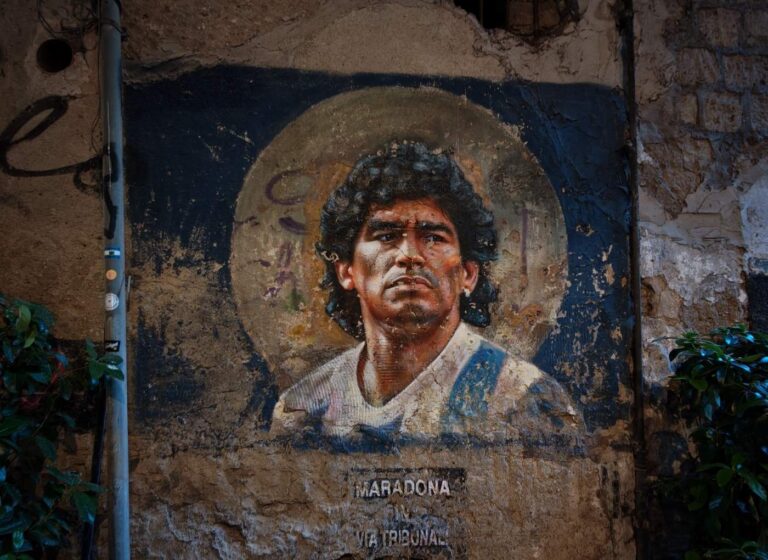 Mural portrait of a man with dark curly hair and a light shirt, with a circular halo behind him on a weathered wall; text 'MARADONA' at the bottom.
