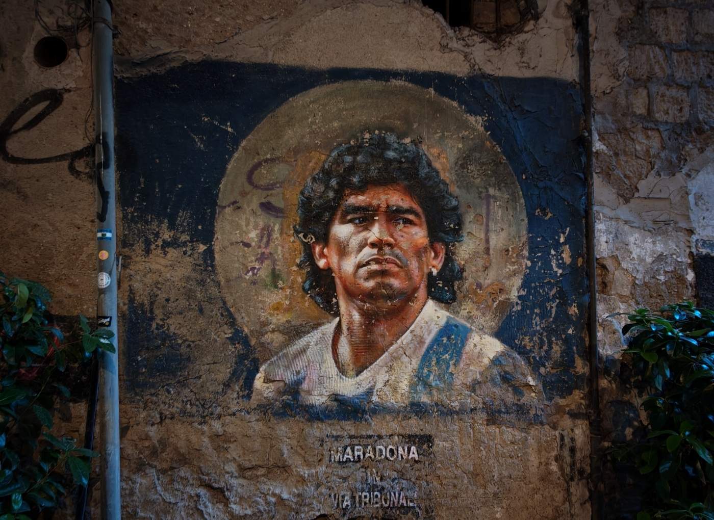Mural portrait of a man with dark curly hair and a light shirt, with a circular halo behind him on a weathered wall; text 'MARADONA' at the bottom.