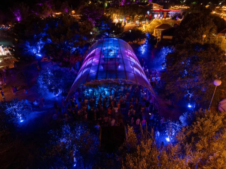 Nighttime outdoor festival under a curved glass canopy lit in blue and purple, with a crowd inside and trees glowing nearby.