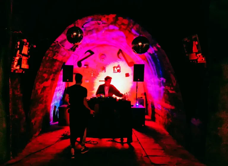 DJ performing at a mixer inside a neon-lit tunnel with pink, red, and purple lighting; two speakers on stands flank the setup and a person stands nearby in silhouette.