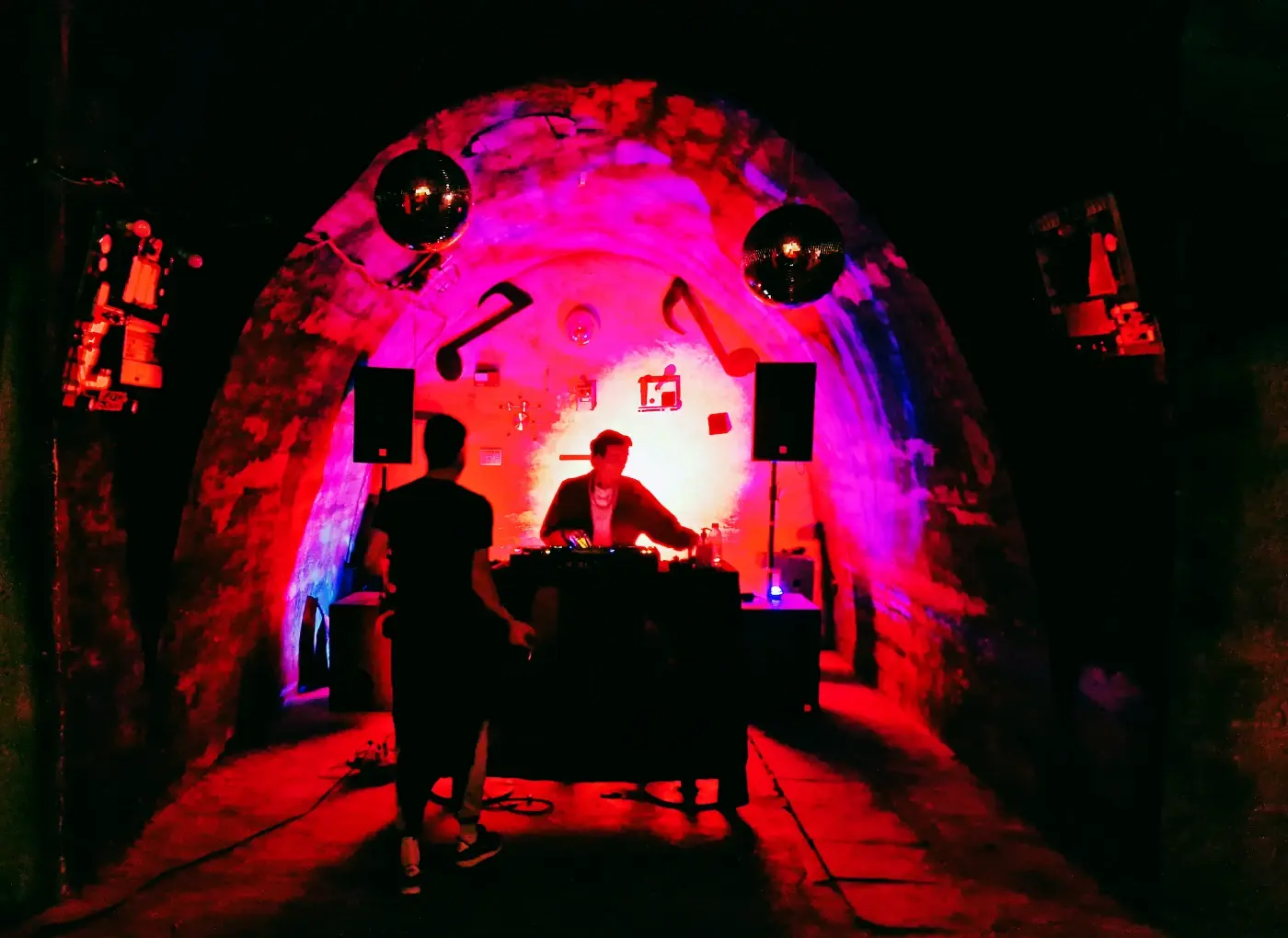 DJ performing at a mixer inside a neon-lit tunnel with pink, red, and purple lighting; two speakers on stands flank the setup and a person stands nearby in silhouette.
