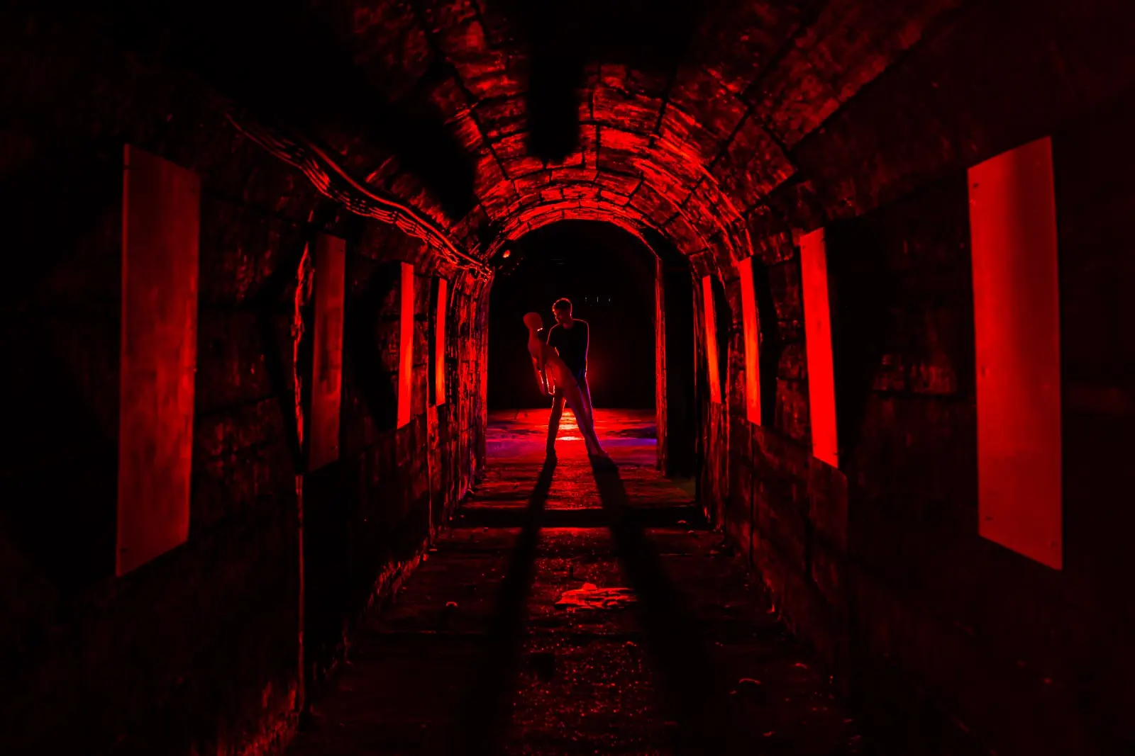 Person stands in a dark, red-lit arched tunnel, cradling a life-size mannequin/dummy.