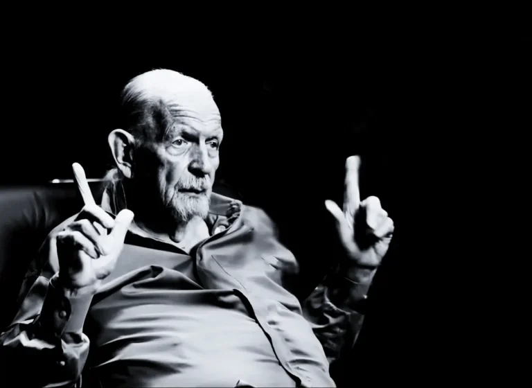 Black-and-white portrait of an elderly man with a white beard, gesturing with both hands, index fingers raised as if signaling.